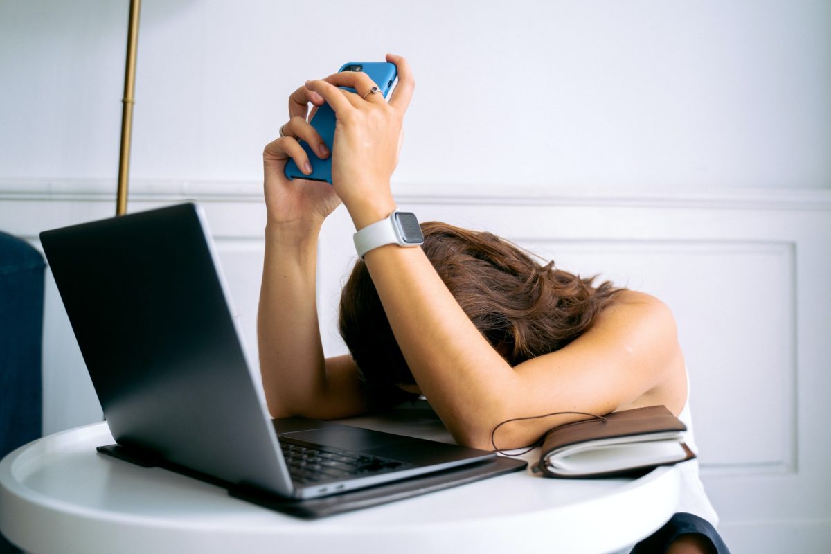 Woman in front of computer with her head down and holding cell phone over her head, indicating frustration