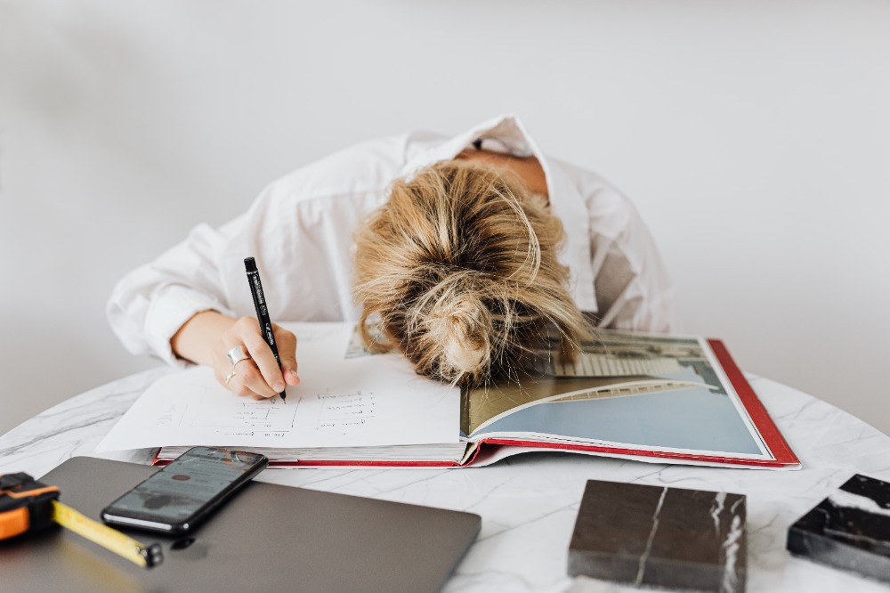 Woman with her head down on a large book holding a pen suggesting exhaustion or frustration.