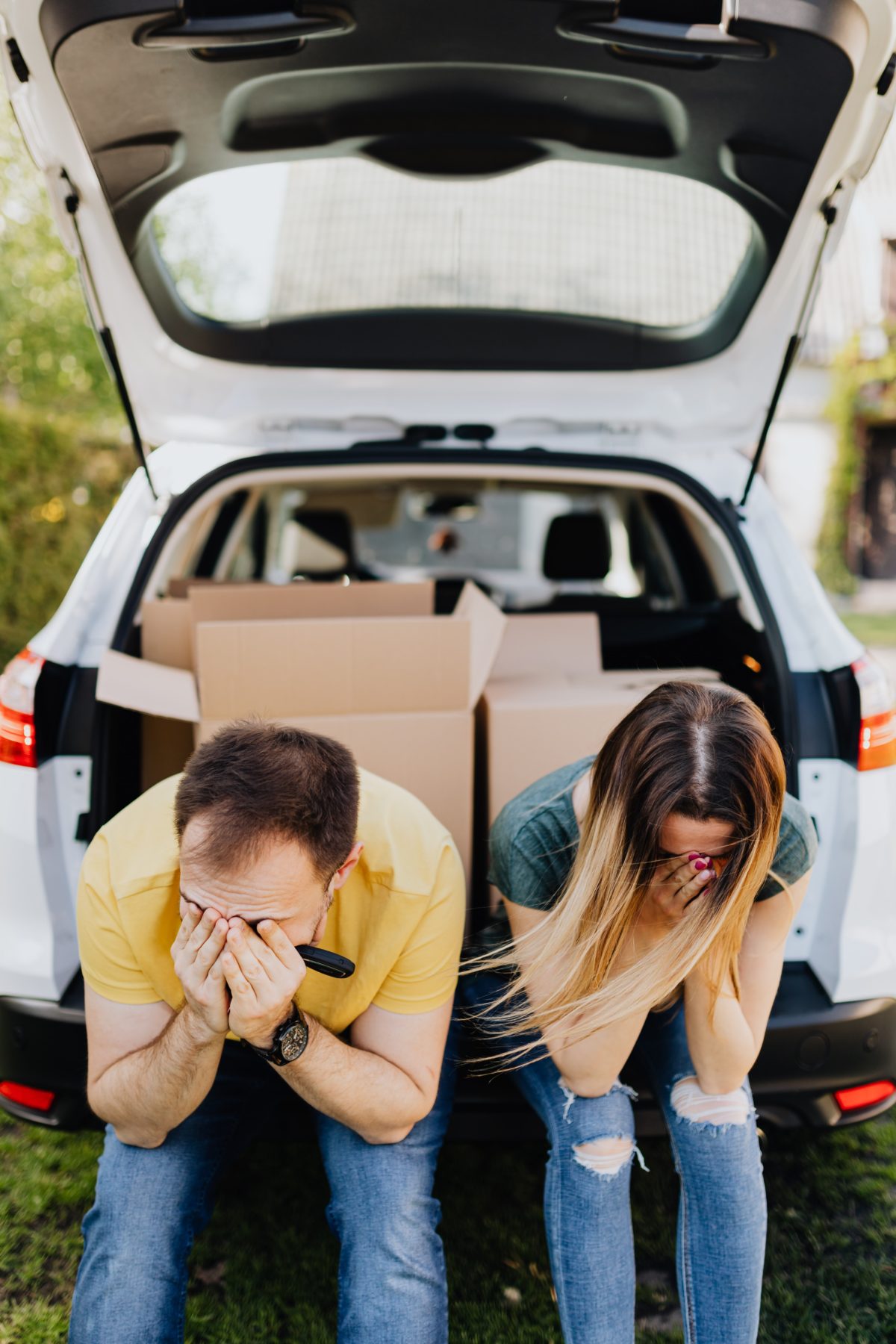 Couple sitting on the back of their vehicle with their heads down and boxes behind them.