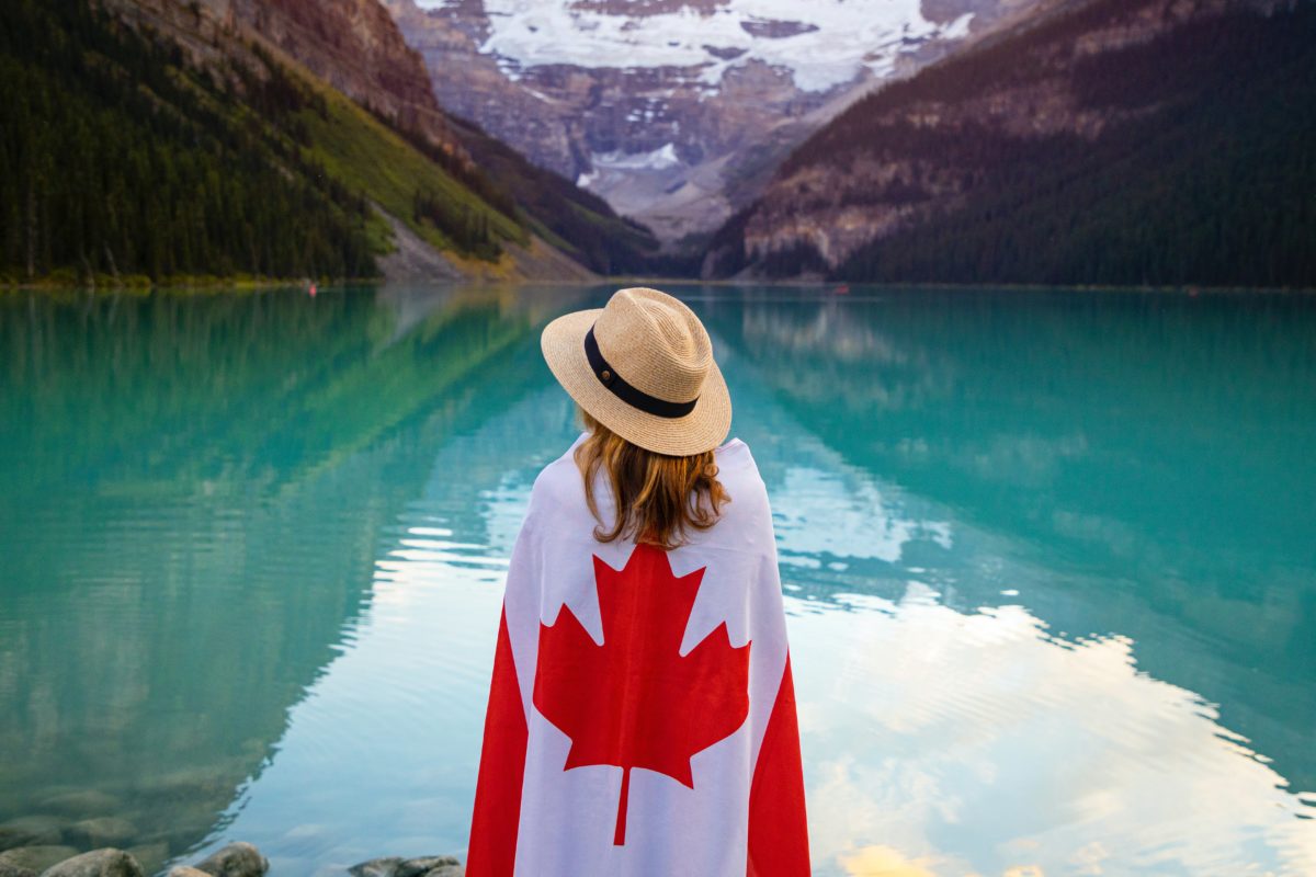 Woman wearing Canadian flag facing away from camera towards emerald lake and snowcapped mountain.