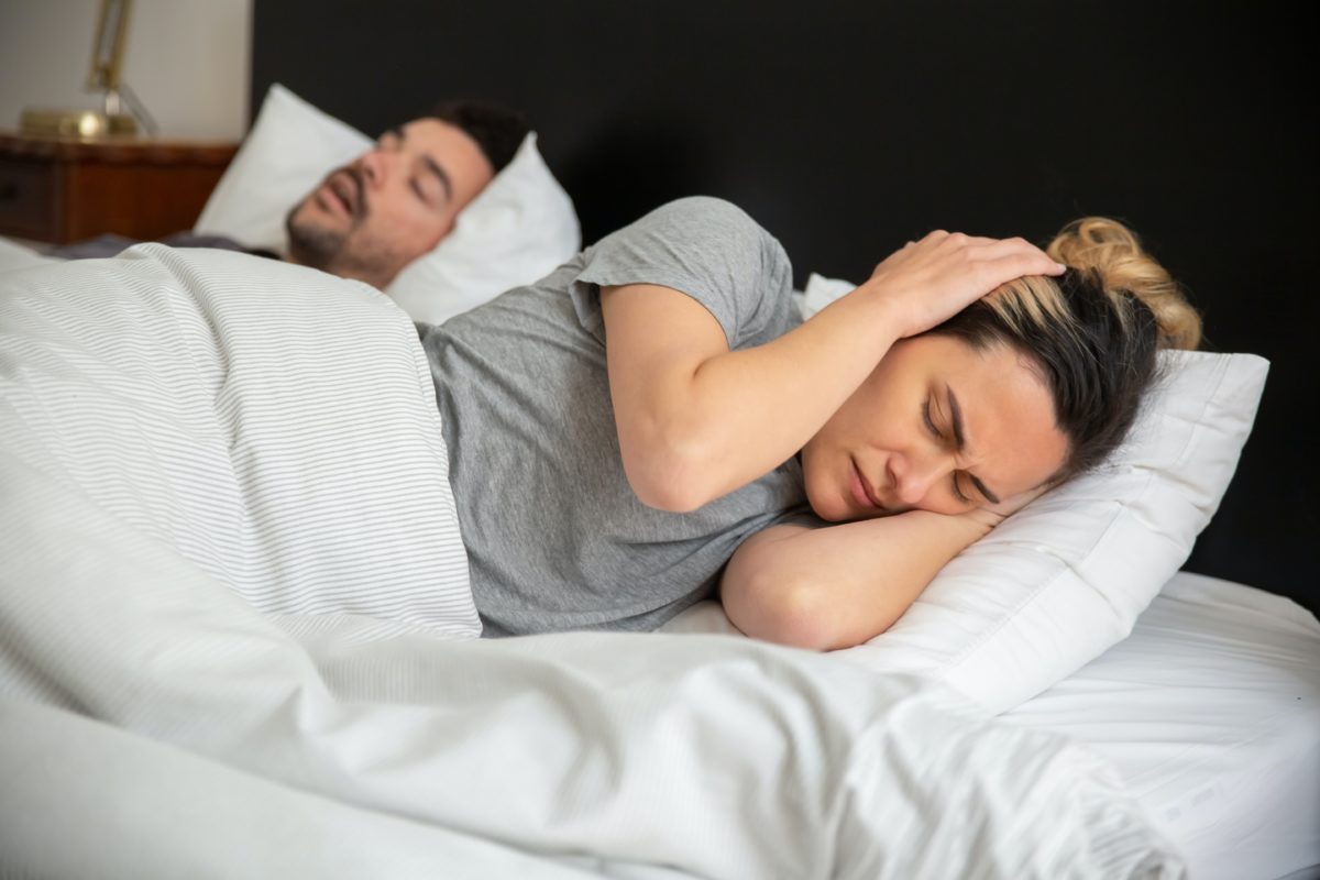 Woman holding her ears sleeping next to man snoring.