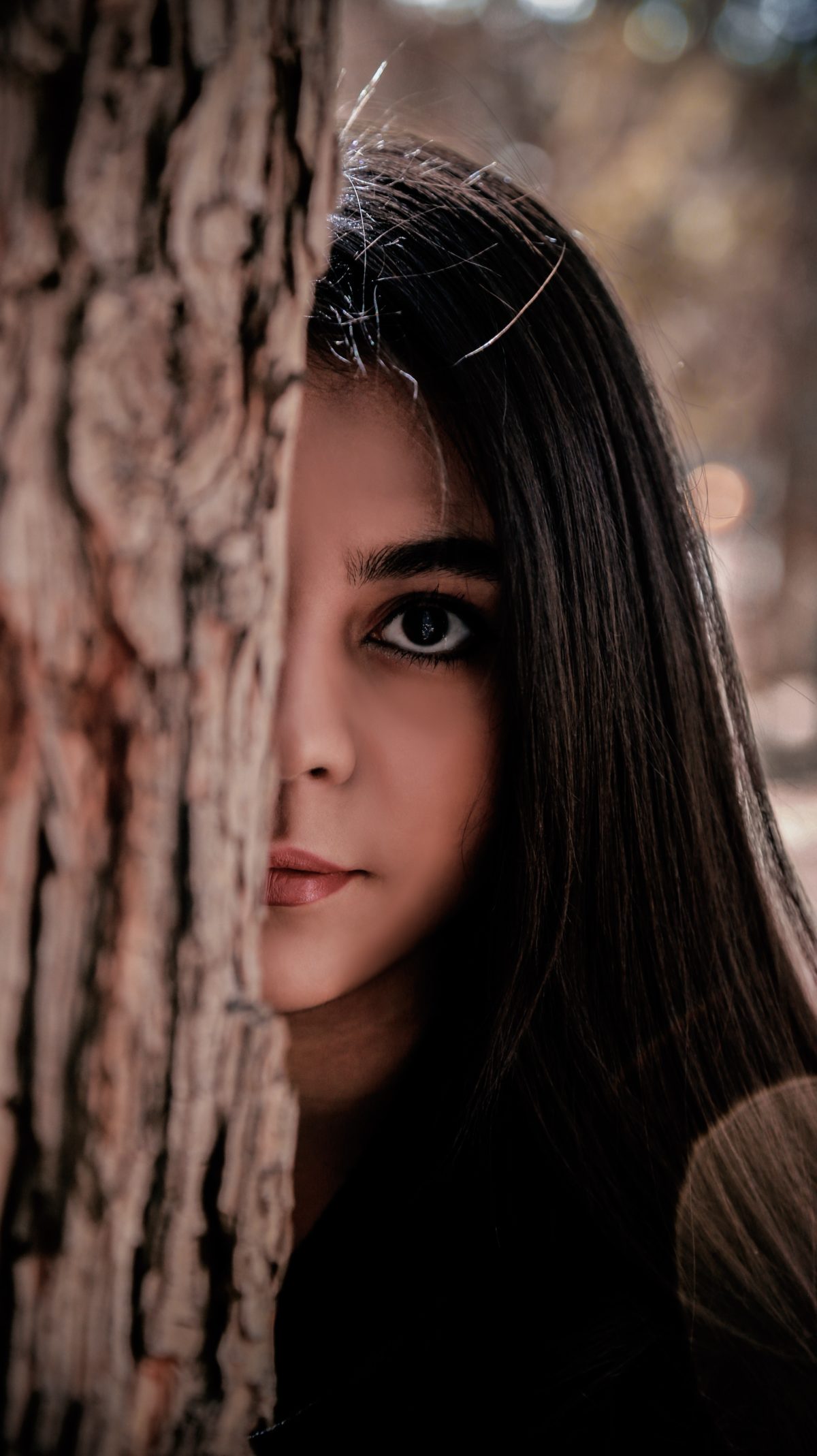 Serious looking young woman whose face is partially obscured by a tree trunk.