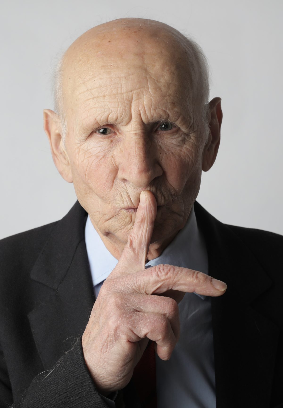 An older man holding a finger up to his lips to make the silence gesture.