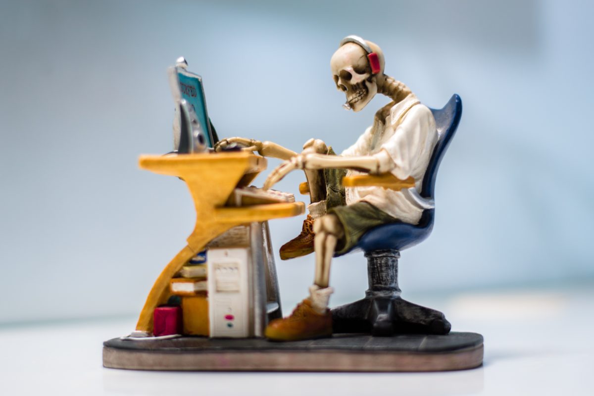 A fully clothed skeleton sitting at a desk and typing at a computer.