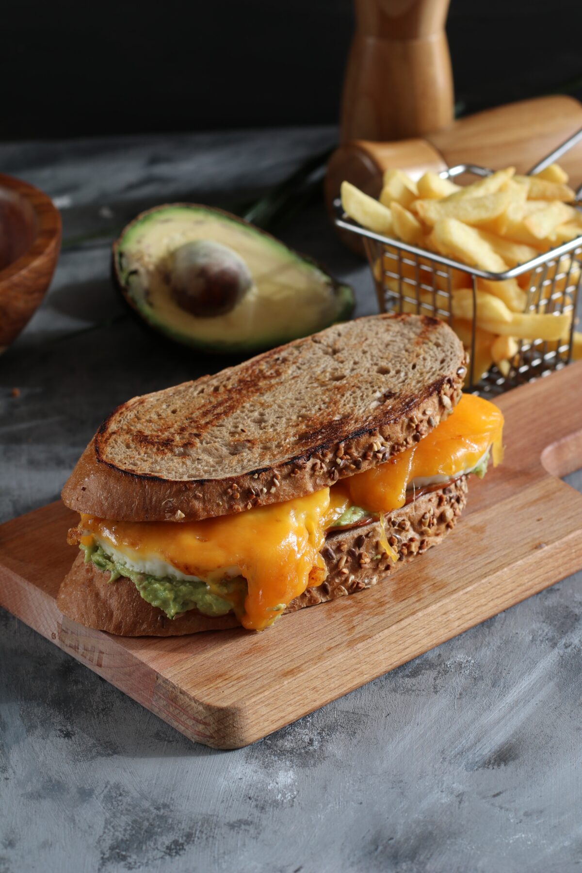 Grilled cheese sandwich on a board with avocado and fries in the background.