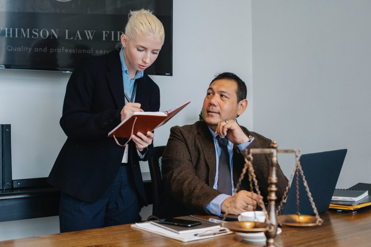 A lawyer looking at a book while standing over another lawyer who is seated at his desk.
