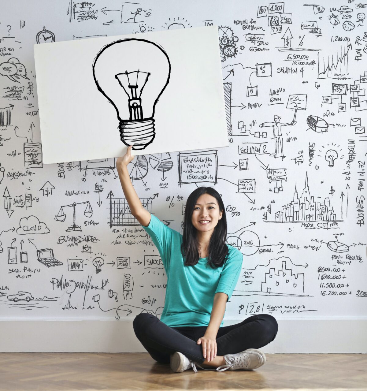 Woman holding up a picture of a light bulb against a background of a wall-board full of diagrams.
