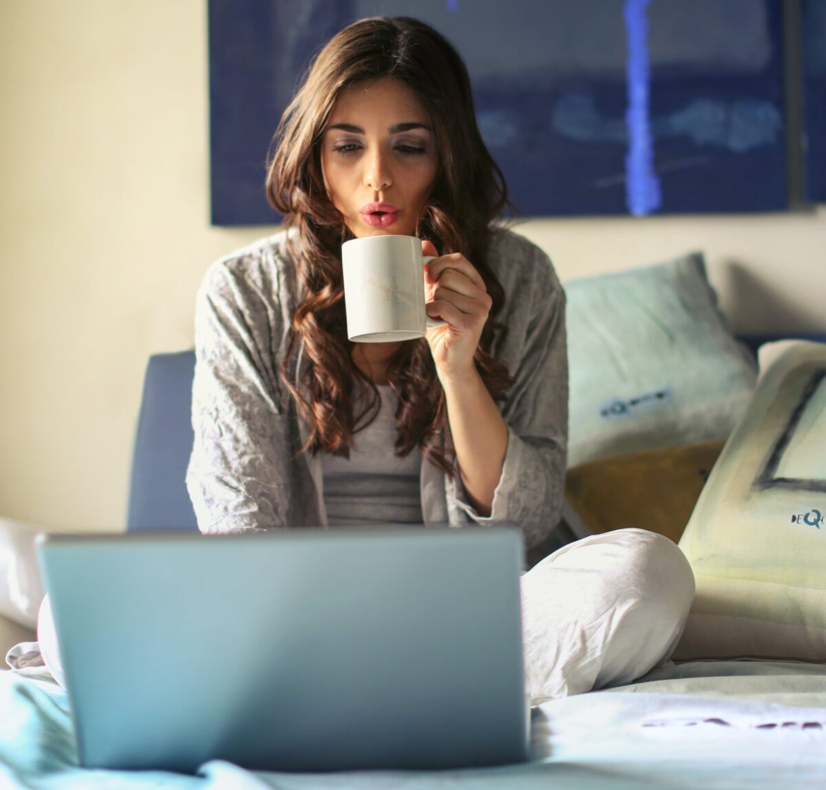 Woman sitting on bed, drinking from mug, and looking at her computer.