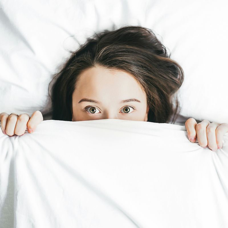Young woman pulling the covers up to her eyes and looking frightened.