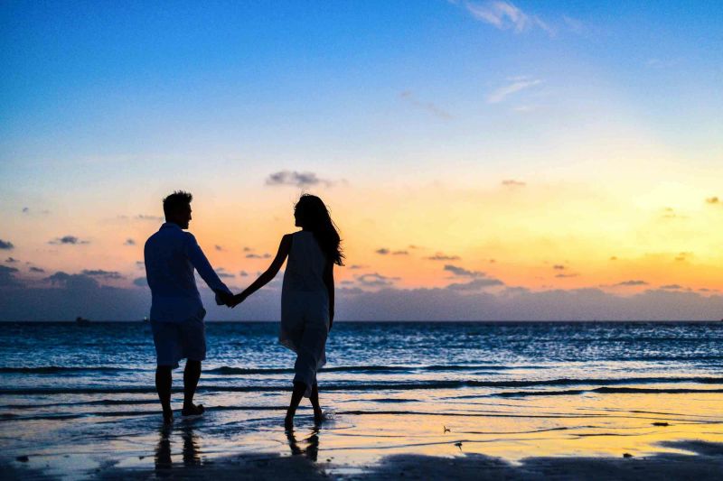 A couple holding hands on the beach with the sun setting behind them.