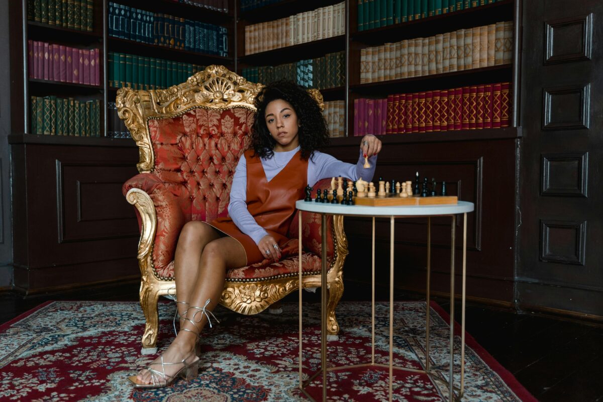 Woman sitting in a gilded chair in a library with a chess set, looking rich and intelligent.