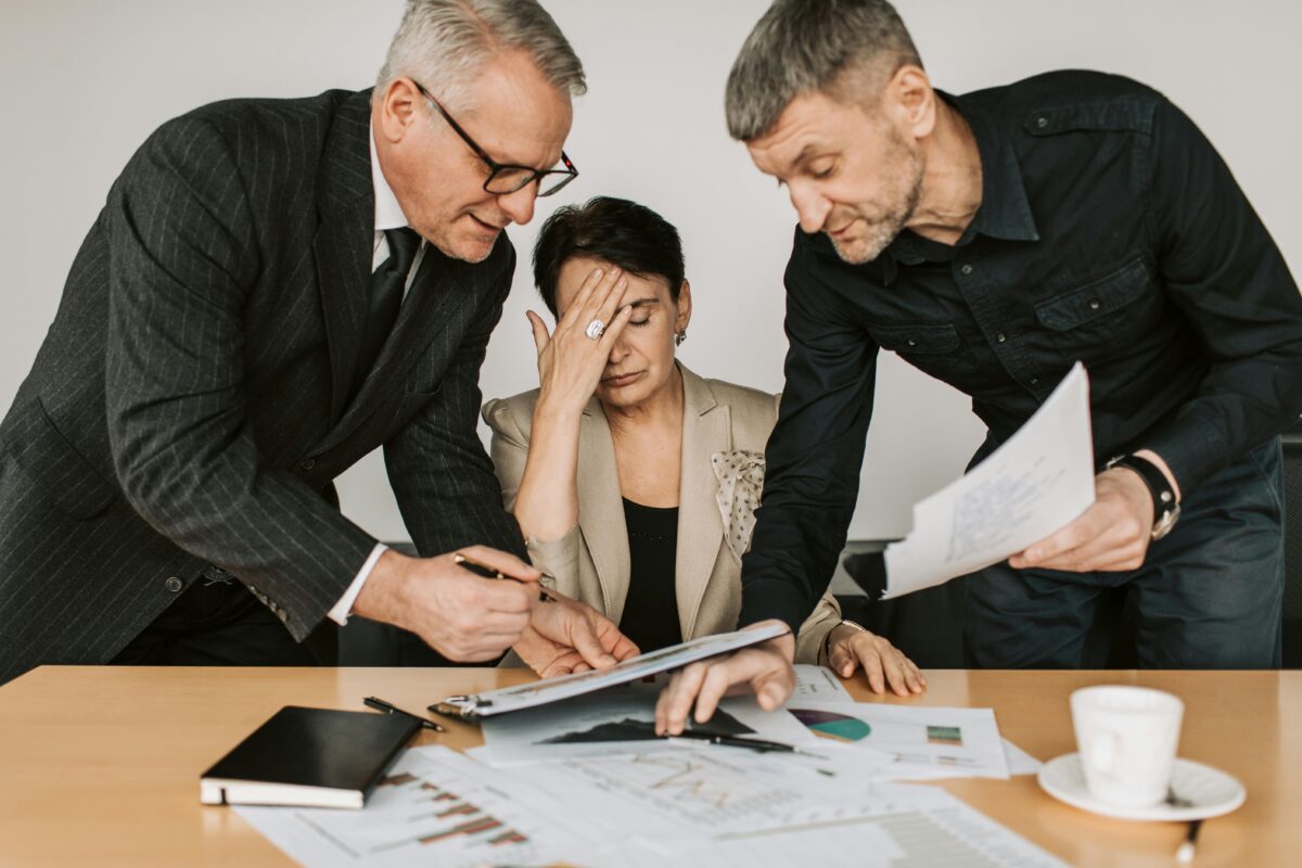 Two men arguing at a business meeting with a woman looking frustrated and stressed.