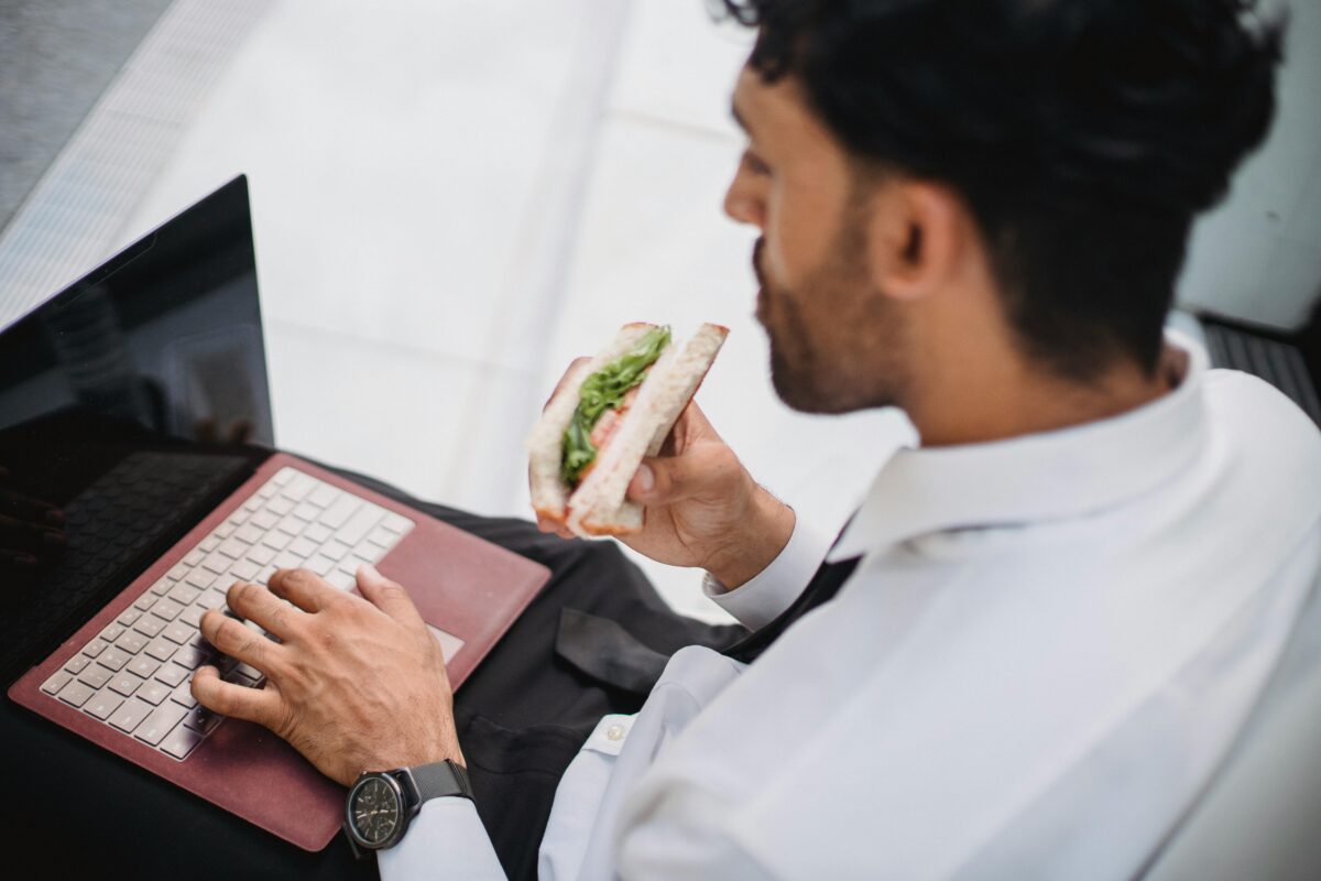 A man eating a sandwich while working on a computer.
