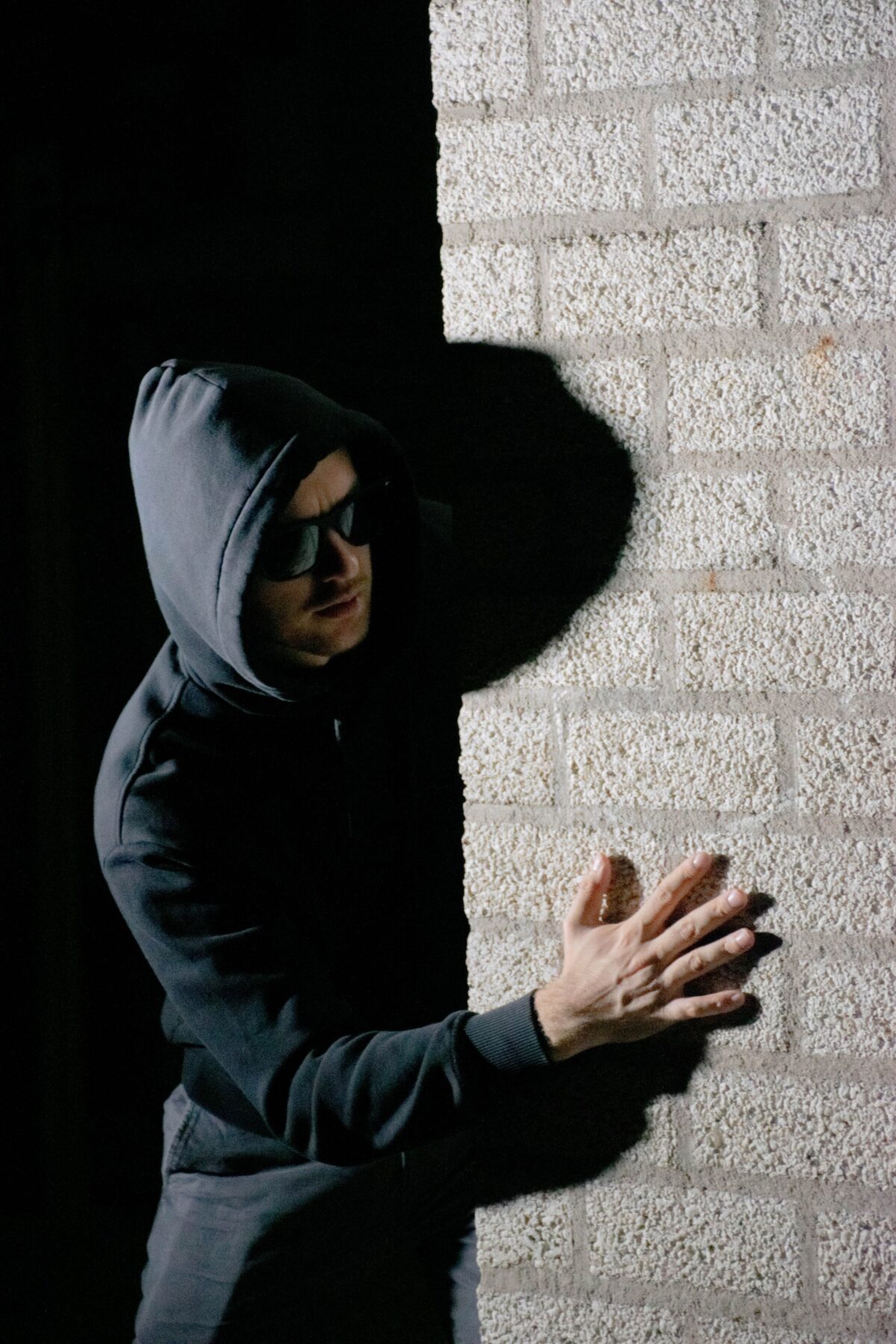 A man in a dark coat, hoody and glasses sneaking around a corner wall.